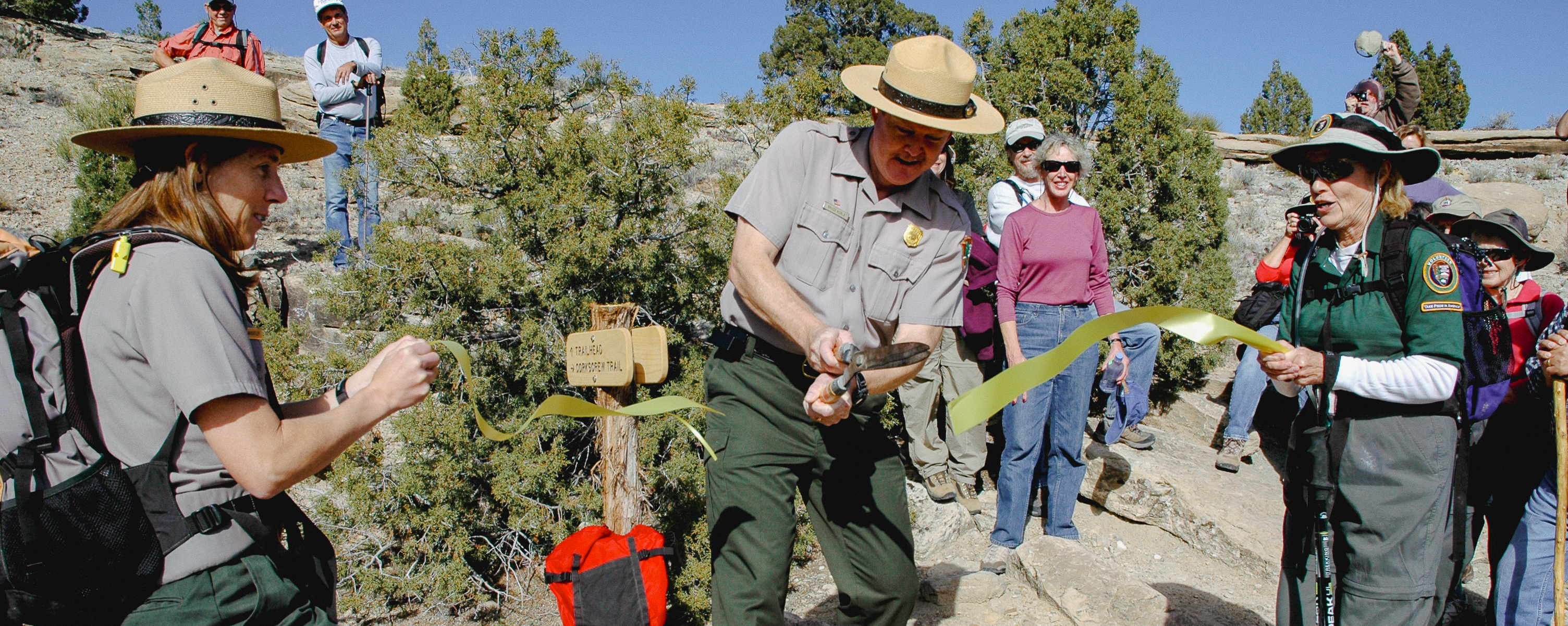 two rangers hold a yellow ribbon as a third ranger cuts it in half while visitors stand in background