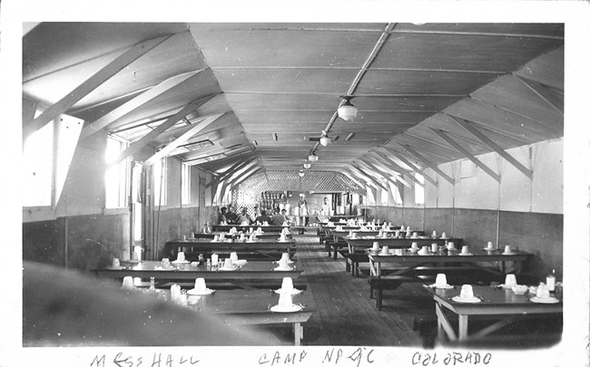 Room with 10 wooden tables and benches in two rows. 6 table settings with plate, silverware and coffee cup at each table. Men are seated at the far table.