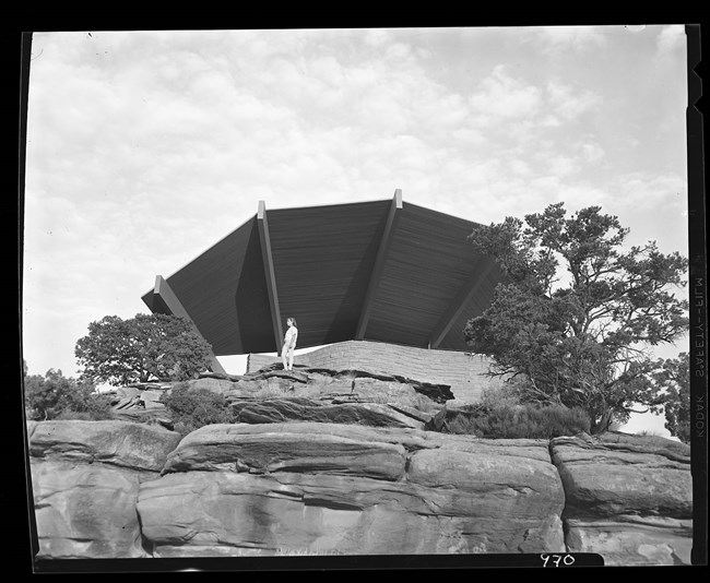 Looking up a rock face, a young woman stands looking out from underneath a cantilevered roof fanning out above her.