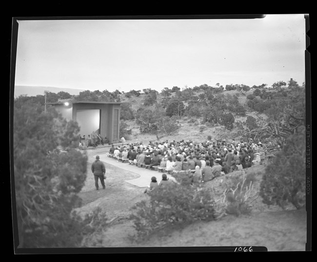 Black and white image of people sitting outside on wooden benches watching a presentation in front of a large building with a screen.