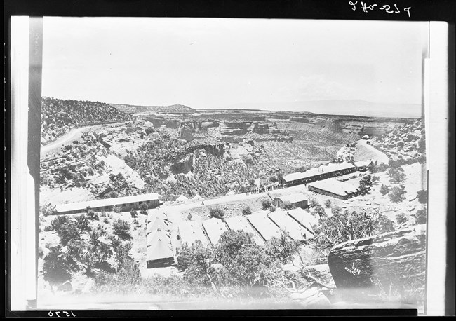 Black & white photo looking down a hillside at a variety of buildings along a road. Men are standing along the road. Canyons are in background.