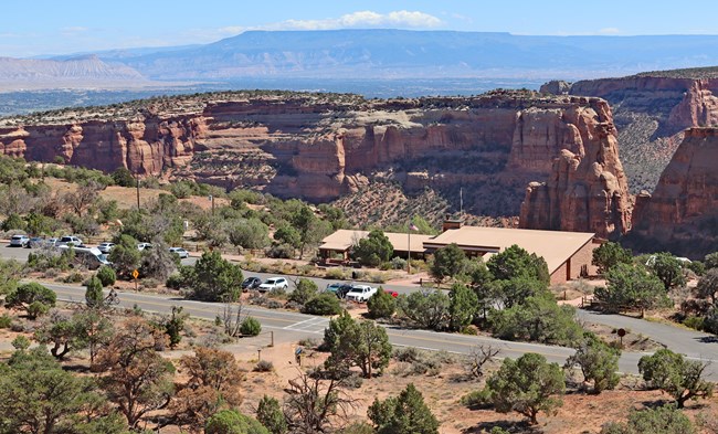 Photograph is looking down at a parking lot and the tan roof of a sandstone brick building. Beyond the building is a canyon and in the distance is a large flat-topped mountain.