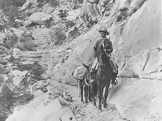 John Otto riding his horse and leading a pack burro, along a rocky trail.