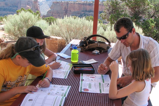 man and his three daughters work on paper booklets at outdoor picnic table