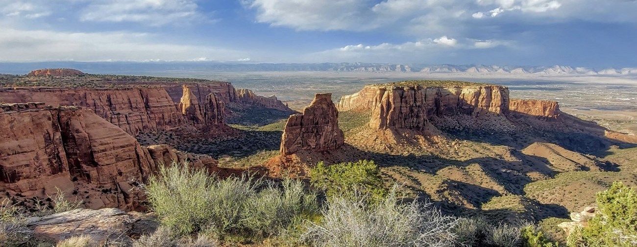Towers of red-orange sandstone cliffs frame a wide canyon with greenery at the bottom and tops.