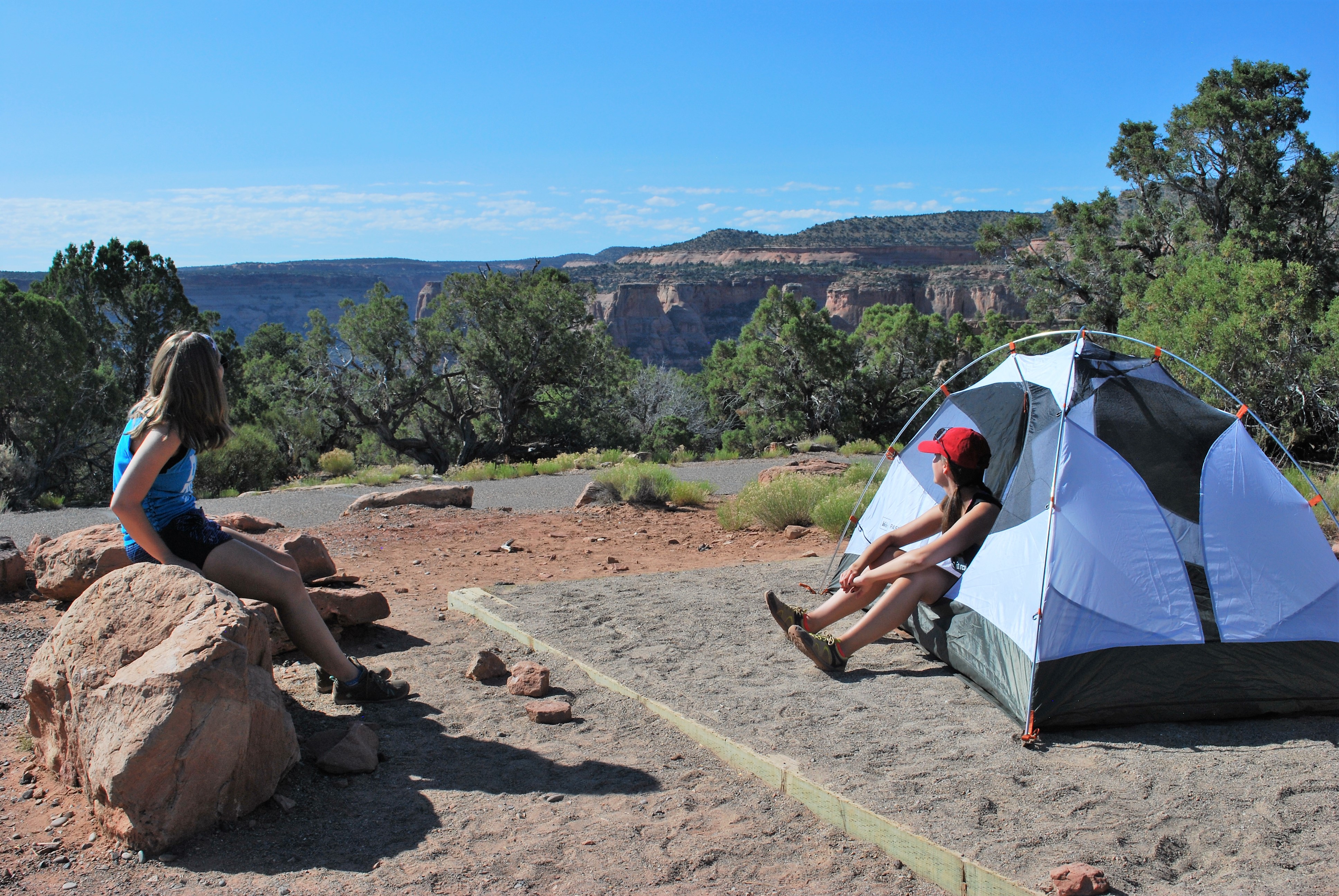 Two female campers sit at a campsite overlooking a orange-tan canyon filled with juniper trees and sheer cliffs.