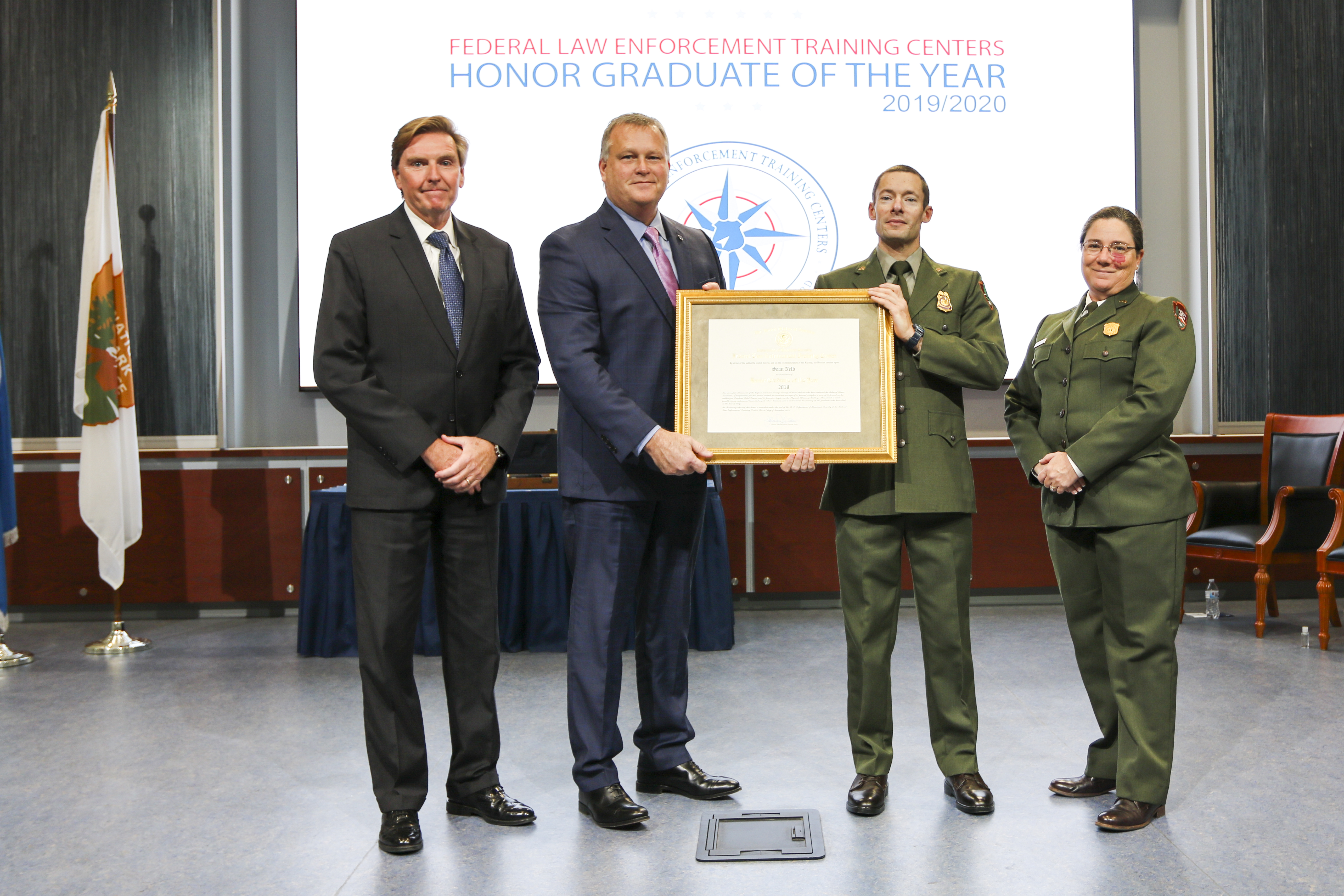 Two men in suits and a man and woman in ranger uniforms standing in a line facing the camera. The center man in the suit and man in uniform holding a framed plaque.
