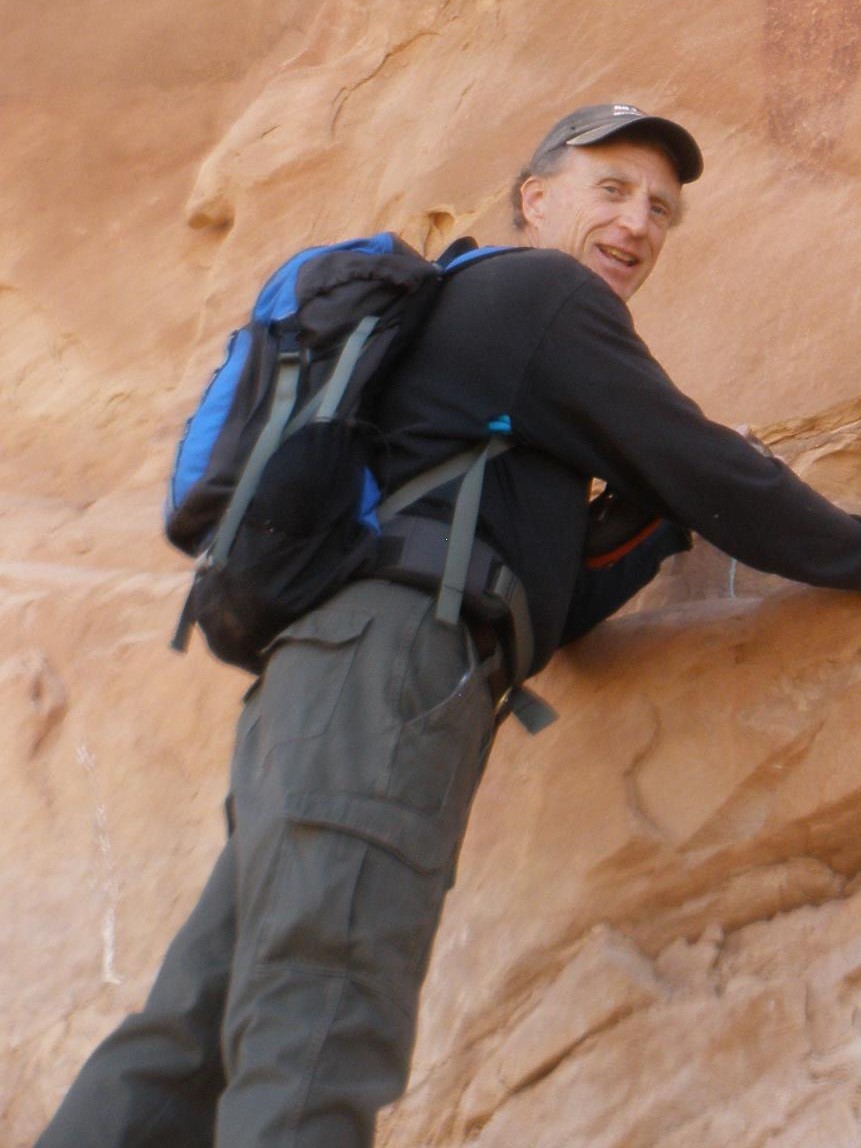 66 year old white male dressed in dark clothes with a blue pack standing against a tan rock wall.