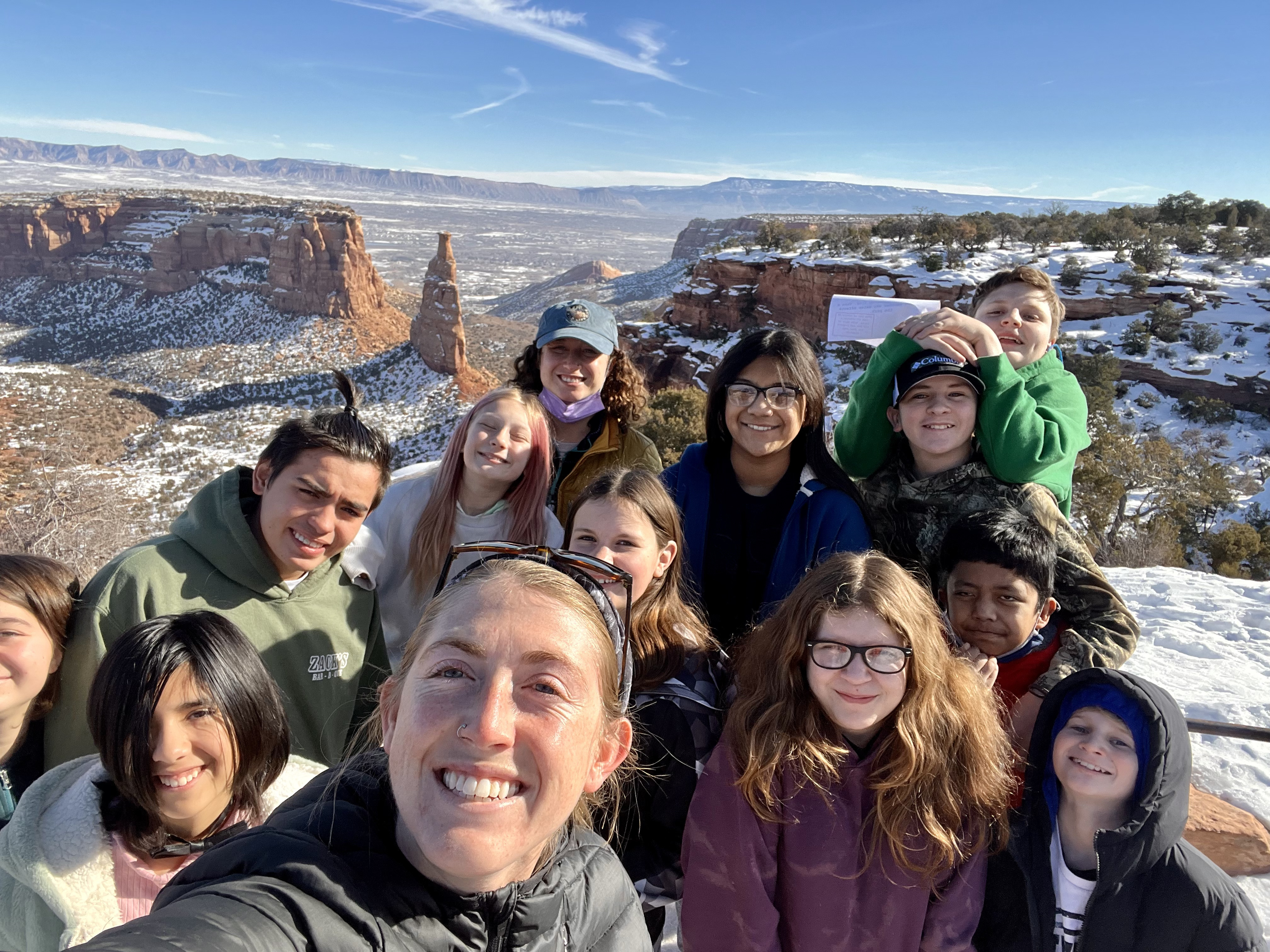 REC Selfie Approved Use Ind. Mon Overlook Group of 12 students and one adult looking up at camera with a canyon with large rock monolith in near background. Far background includes a distant escarpment and immense flat-topped mountain.