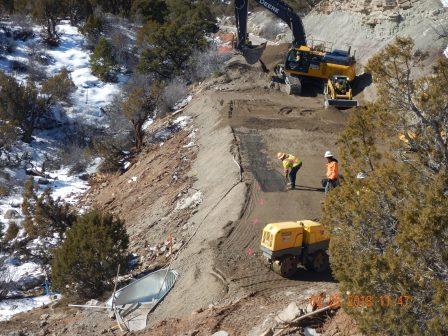 Construction crew lays geo-fabric while heavy equipment operators wait to lay dirt over it. A compactor is ready to compact the new fill.