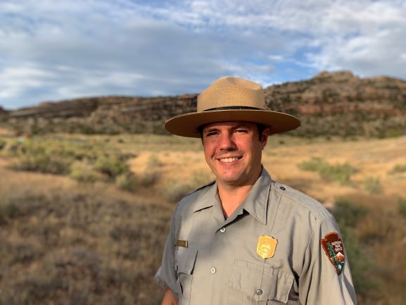 Nathan Sounder in Park Service uniform standing outside. Behind him is tan grasses and rock outcrop.