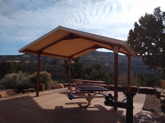 Two covered picnic tables with views of the cliffs and monoliths, and a juniper tree to the right of the tables.