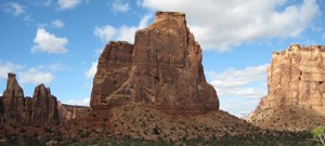 Independence Monument, a wide tower of red-orange sandstone, stands among other similar towers in a wide, green canyon