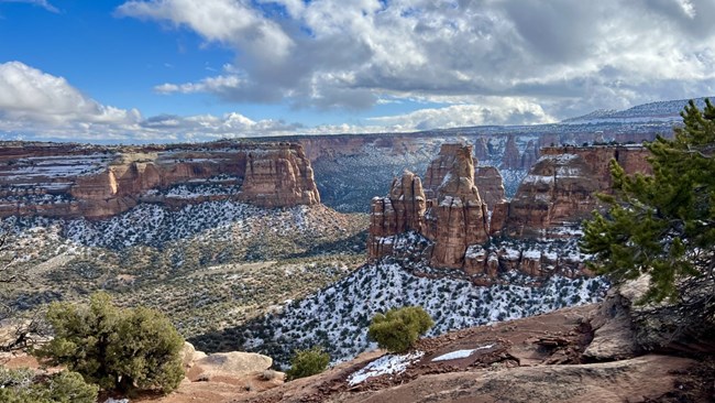 Red-rock cliffs and towers, sparse green shrubs and trees, blue sky with fluffy white clouds, recent snowfall covering the shadows