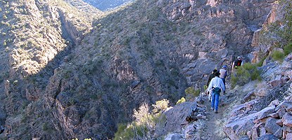 Four hikers along Corkscrew Trail's steep edge of forested canyon hills of dark gray rocks