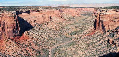 Sheer walls of red-orange sandstone frame a wide, flat, green canyon bottom. A dry creekbed winds through.