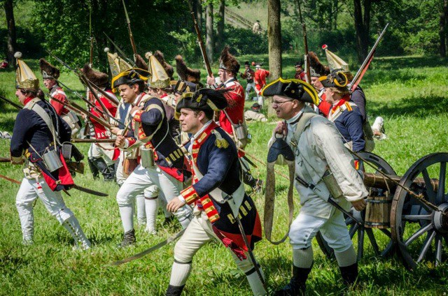 Reenactors dressed as British royal troops march through tall grass.