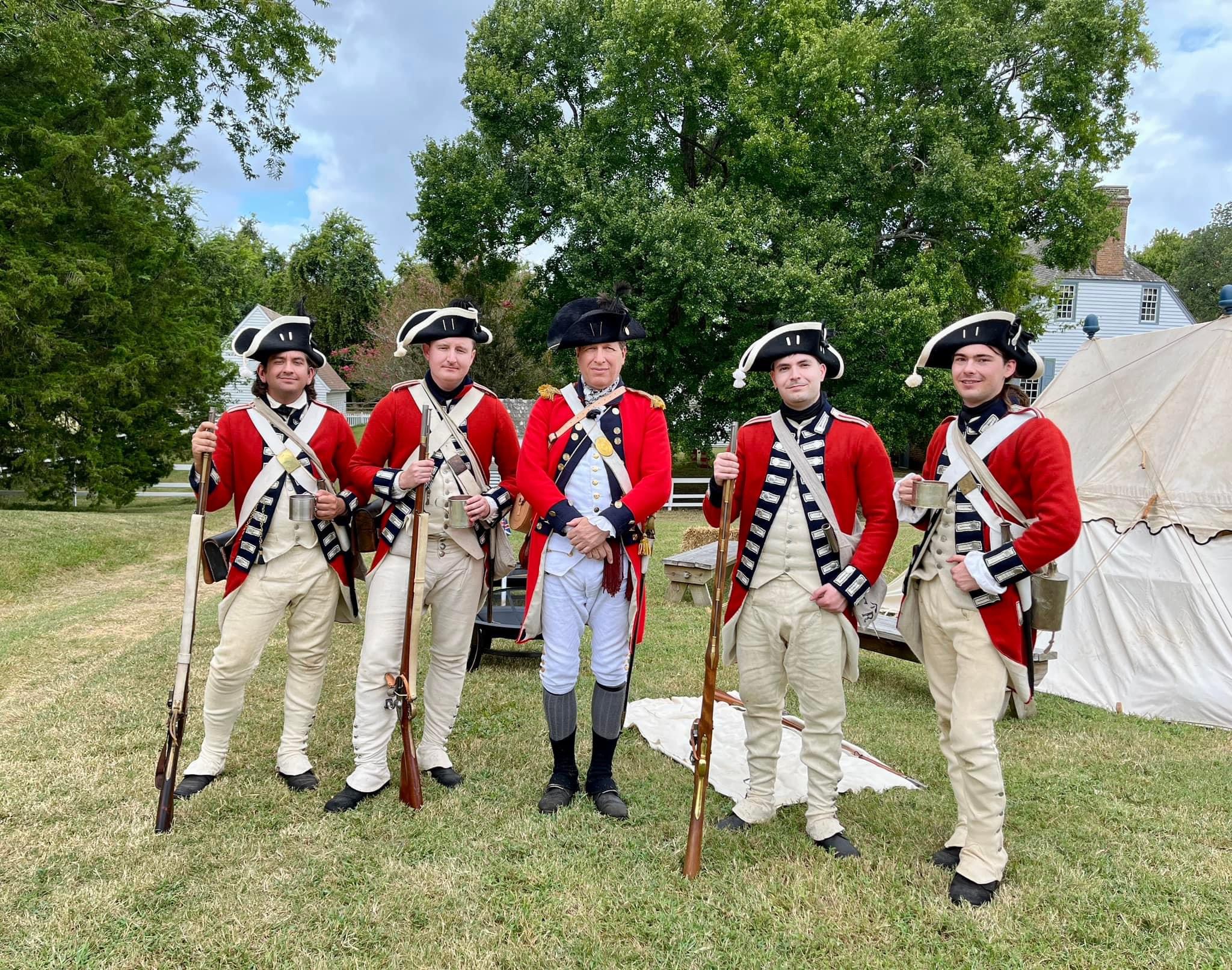 A group of five historical reenactors dressed as British soldiers. Three of the men have muskets. the  fourth is holding a cup. In the center is an officer.