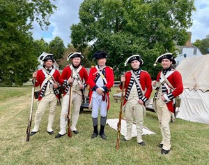 A group of five historical reenactors dressed as British soldiers. Three of the men have muskets. the fourth is holding a cup. In the center is an officer.