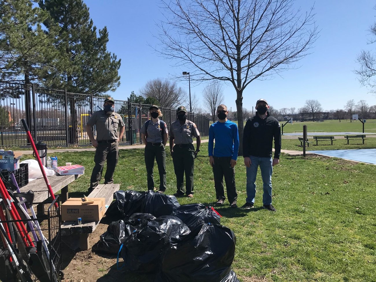 Three Rangers, the Coltsville Community Volunteer Ambassador, and a volunteer stand together looking at the camera. In front of them is a pile of full trashbags and tools for picking up trash. They are in a green field in Colt Park, Hartford, CT.