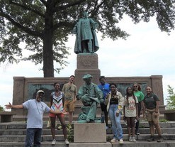 A group of smiling people standing on the steps of a monument surrounding the lower statue of a man sitting with his leg crossed on his knee.