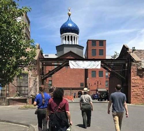 Image of a blue onion dome atop brick buildings with a ranger leading five people towards the buildings. Their bacls are to the camera