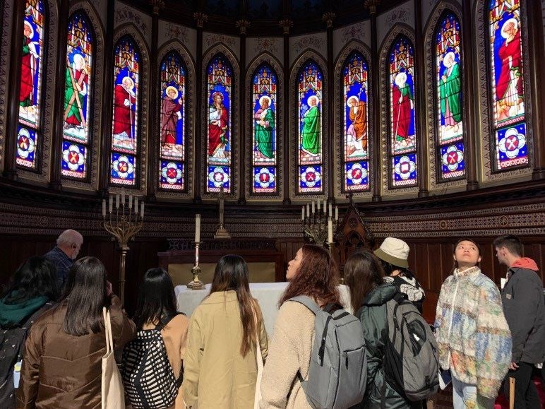 Seven students with their backs to the camera viewing 11 stained glass windows of the apostles of Jesus.