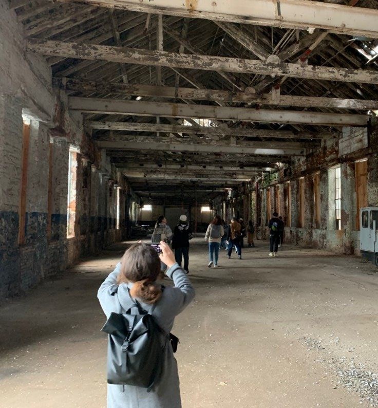 A student with their back to the camera holds a camera to take photos inside an empty facotry building. A group of students are in front of them.