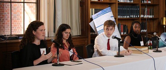 Four students sitting at a table facing an audience. There are microphones on the table and one of the students is speaking into a microphone to the audience that is not shown.
