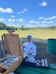 a smiling young woman working from a easel as she painted the surrounding grassy park scenepark