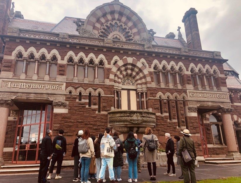 A group of students with a ranger with their backs to the camera viewing a brownstone building with the words "In Memory of Caldwell Hart Colt" visible on the building.