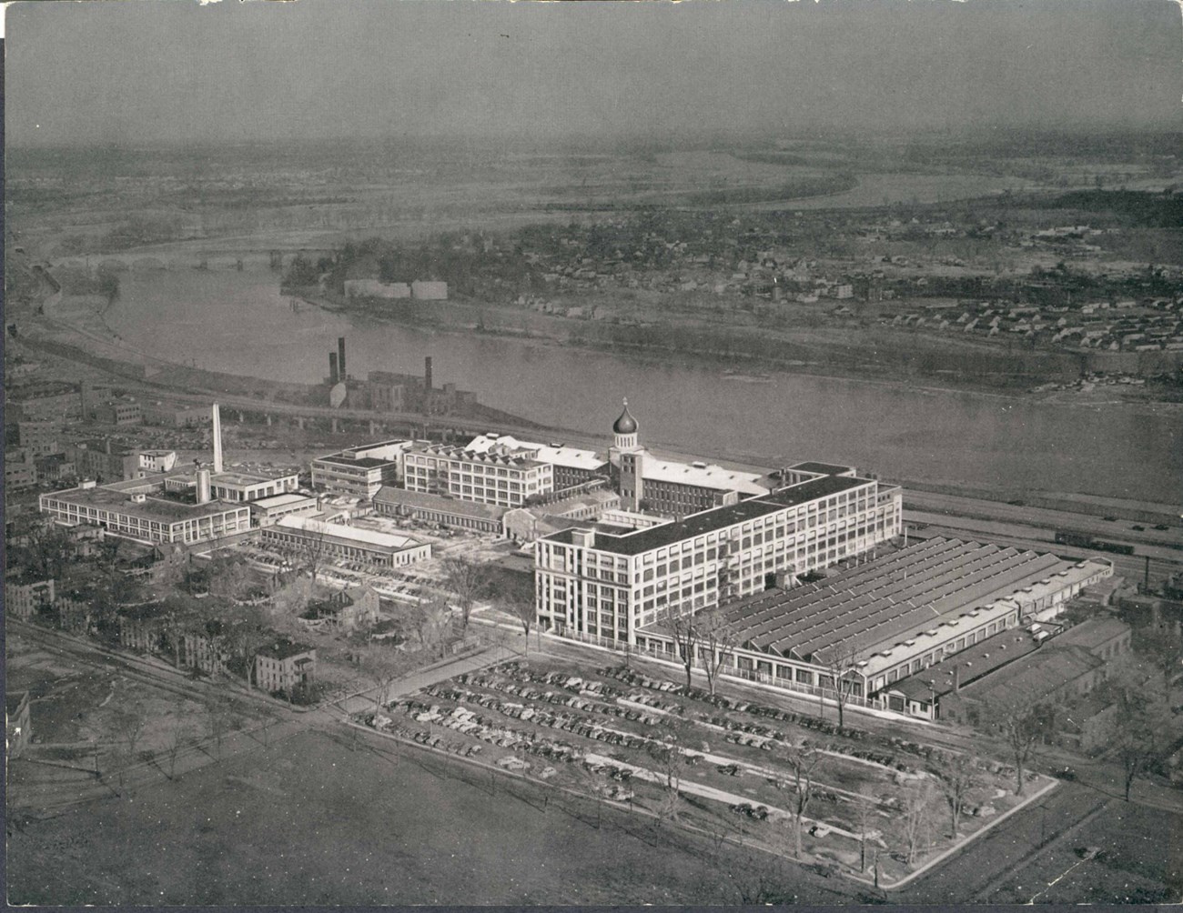 An aerial photo of a factory along a river with houses on the other side of the river.
