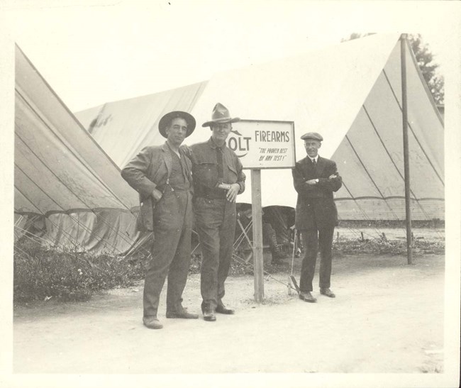 A black and white photo of three men standing next to a sign that reads "Colt Firearms The Proven Best By Any Test!" Tents are behind the men as well.