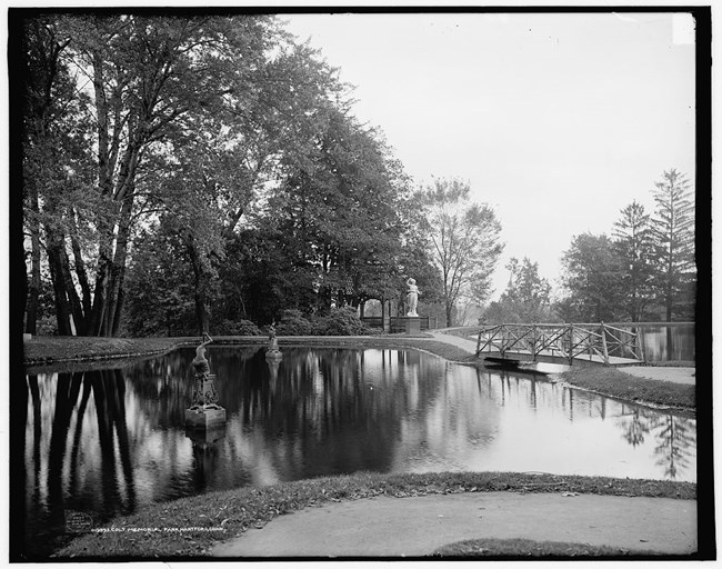 A black and white photo of a pond with a bridge surrounded by statues and trees.