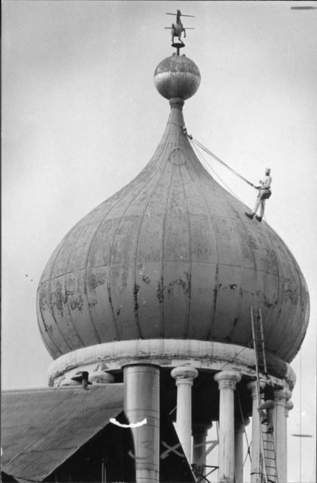 Black and white photo of worker on the Onion Dome being held up by ropes. Another worker is climbing a ladder.