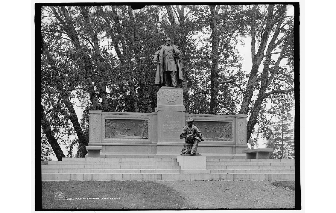 A black and white photo of a memorial statue with to figures, one standing and one sitting and two bronze relief's.