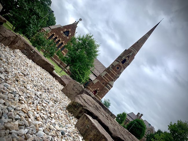 A church with steeple rising towards the sky, behind a memorial made of large stones sitting on small rocks.