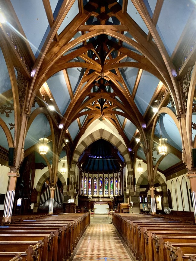 The inside of a church with multiple stained glass windows, wooden pews, stone patterned floors, and wooden arches.