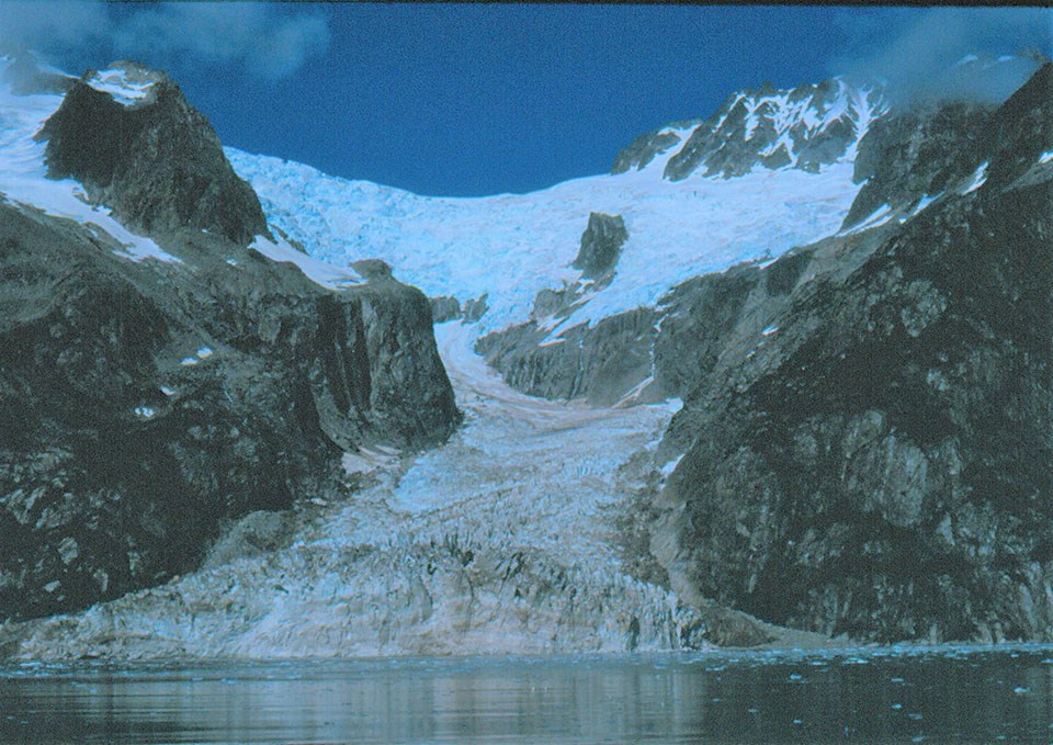 The bottom fifthof the picture is water.  A blue colored glacier flows over a mountain at the top of the picture, and down to the water.  The glacier is narrow in the middle of the image as it flows between two rocky outcroppings.