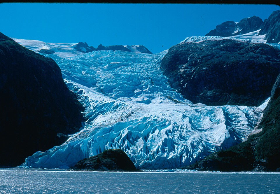 A blue colored glacier flows over a mountainside and down to the water.  There are rocks and land in the water in front of the glacier