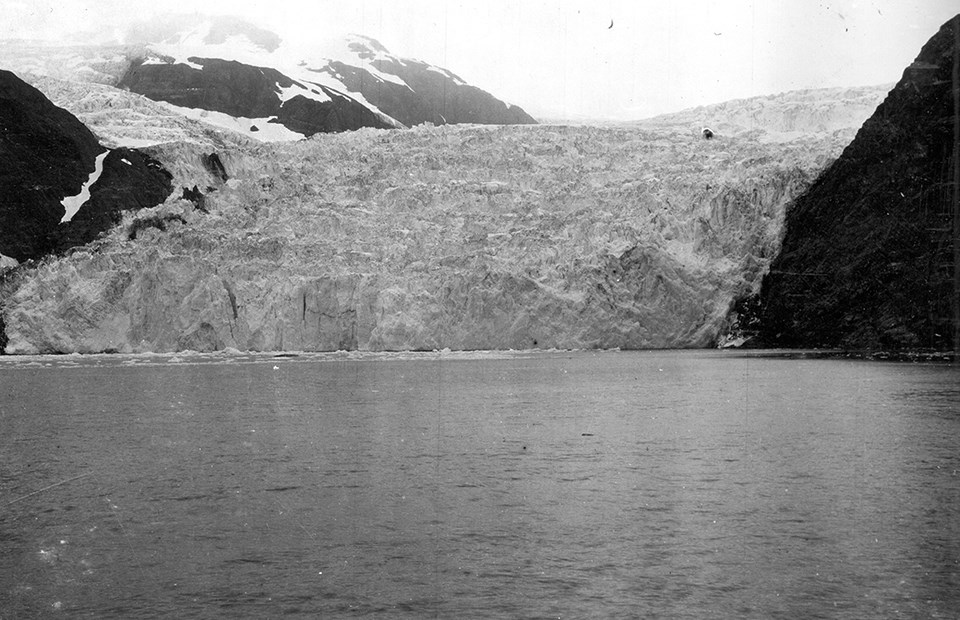 A black and white photo.  The bottom half of the picture is water.  There are mountainsides on the right and left of the picture.  In the middle is a large glacier that meets the water.