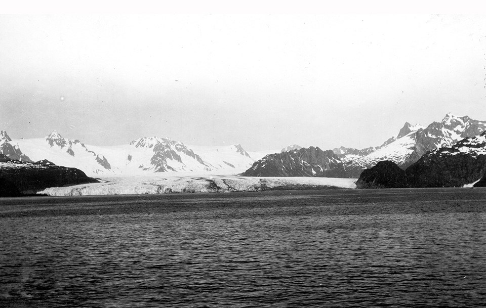A black and white photo.  The bottom half of the picture is water. In the center of the image is a glacier.  On the left and right of hte glacier are rocky outcroppings.  In the background are snow covered mountains