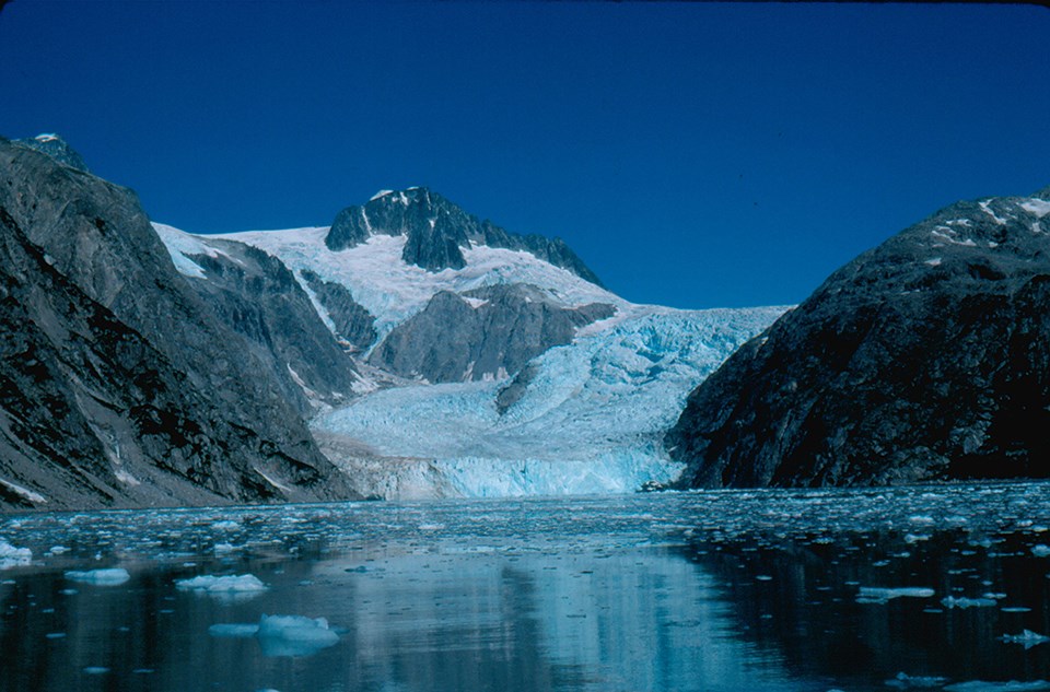 A blueish colored glacier flows from the top center of the image to the bottom center.  Two rocky mountains are on the left of the glacier, and one rounded mountainside is on the right of the glacier.  The bottom third of the image is icy water.