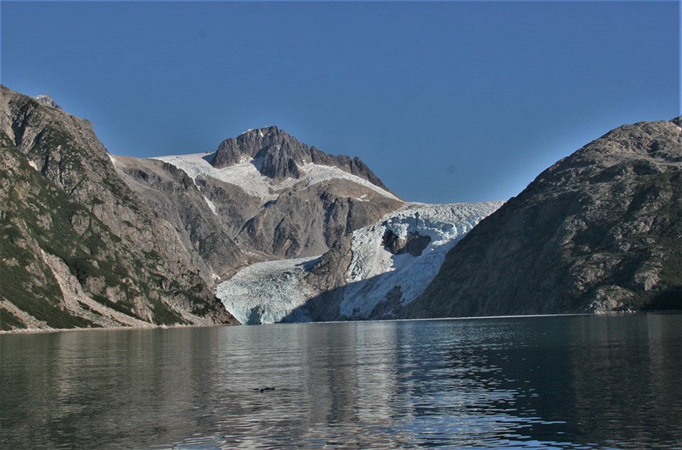 A blueish colored glacier flows from the top center of the image to the bottom center.  Two rocky mountains are on the left of the glacier, and one rounded mountainside is on the right of the glacier.  The bottom third of the image is icy water.