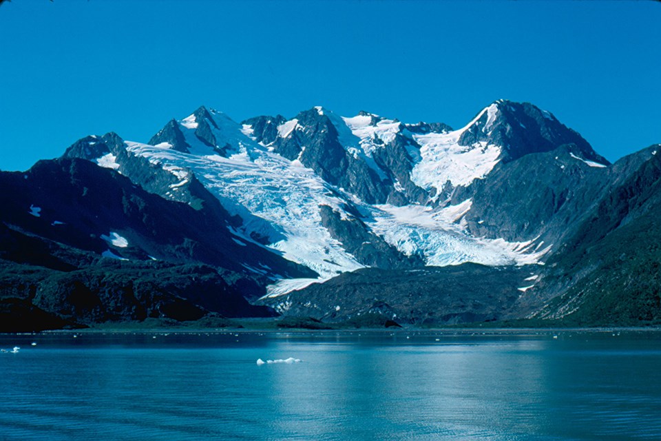 A large mountain is in the center of the image. An alpine glacier flows from the upper left of the mountain down towards the center.  The bottom third of the image is water with ice chunks floating in it.