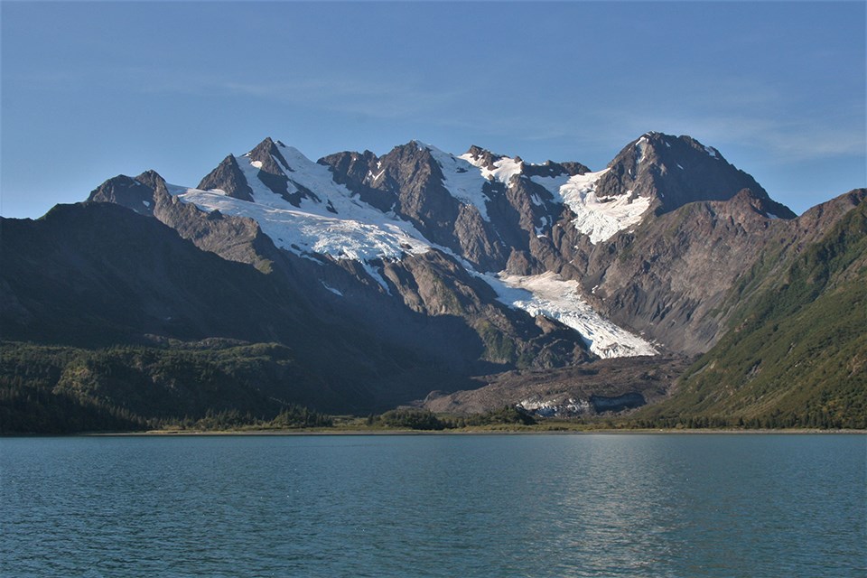 A large mountain is in the center of the image. An alpine glacier flows from the upper left of the mountain down towards the center.  The bottom third of the image is water with ice chunks floating in it.