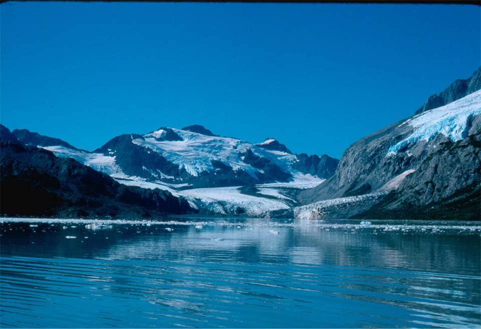 The bottom half of the image is water with chunks of floating ice in it. The top half of the image is mountains with snow patches. In the center of the image a glacier flows from right of center to cent, and down to the water.