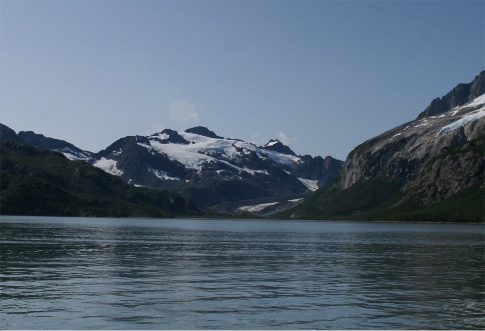 The bottom half of the image is water with chunks of floating ice in it. The top half of the image is mountains with snow patches. In the center of the image a glacier flows from right of center to cent, and down to the water.