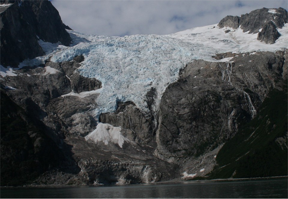 A blue colored glacier flows over a mountainside.  Center right is a large rocky outcropping from undre thglacier.
