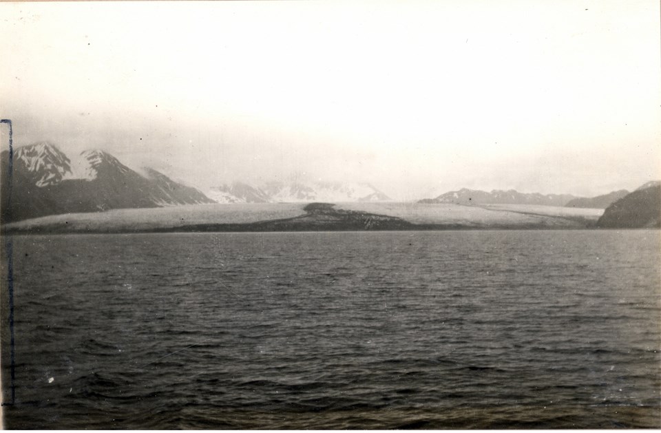 A black and white photo.  The bottom half of the picture is water.  In the top half there is a large glacier in the center of center of the picture.  There are mountains on the right edge and left edge of the picture to the sides of the glacier.